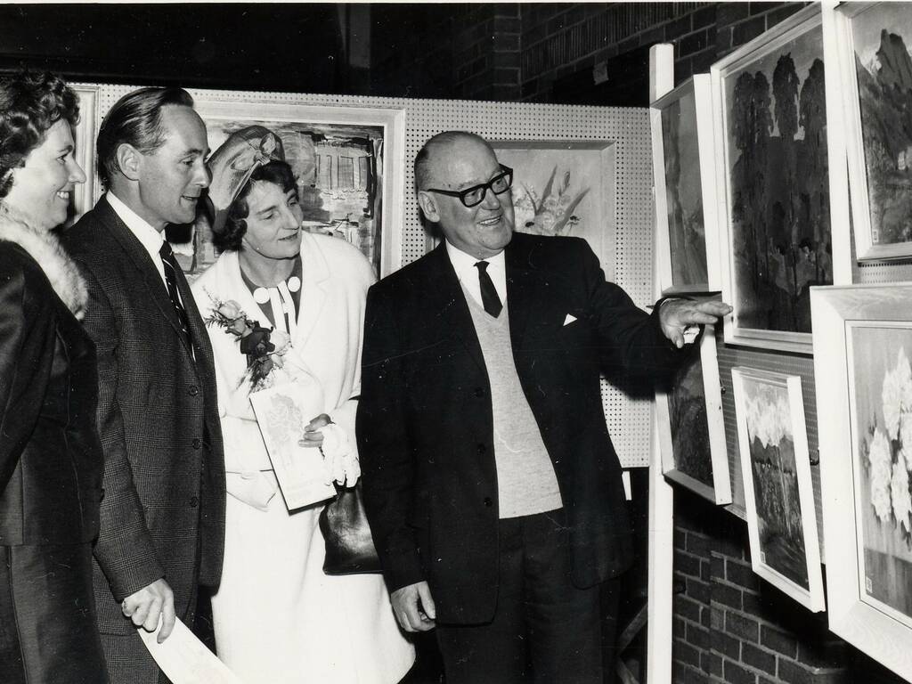 Black and white photo of three individuals being show art pieces by an artist
